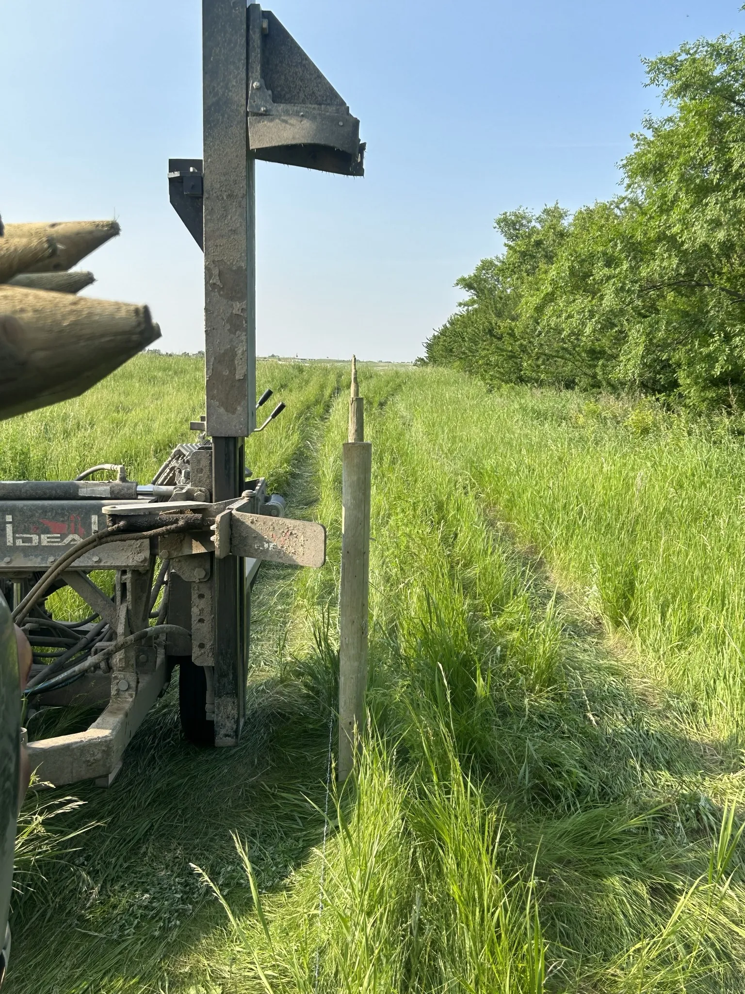 Post pounder driving fence posts along a long prairie fence line through lush green Saskatchewan grassland